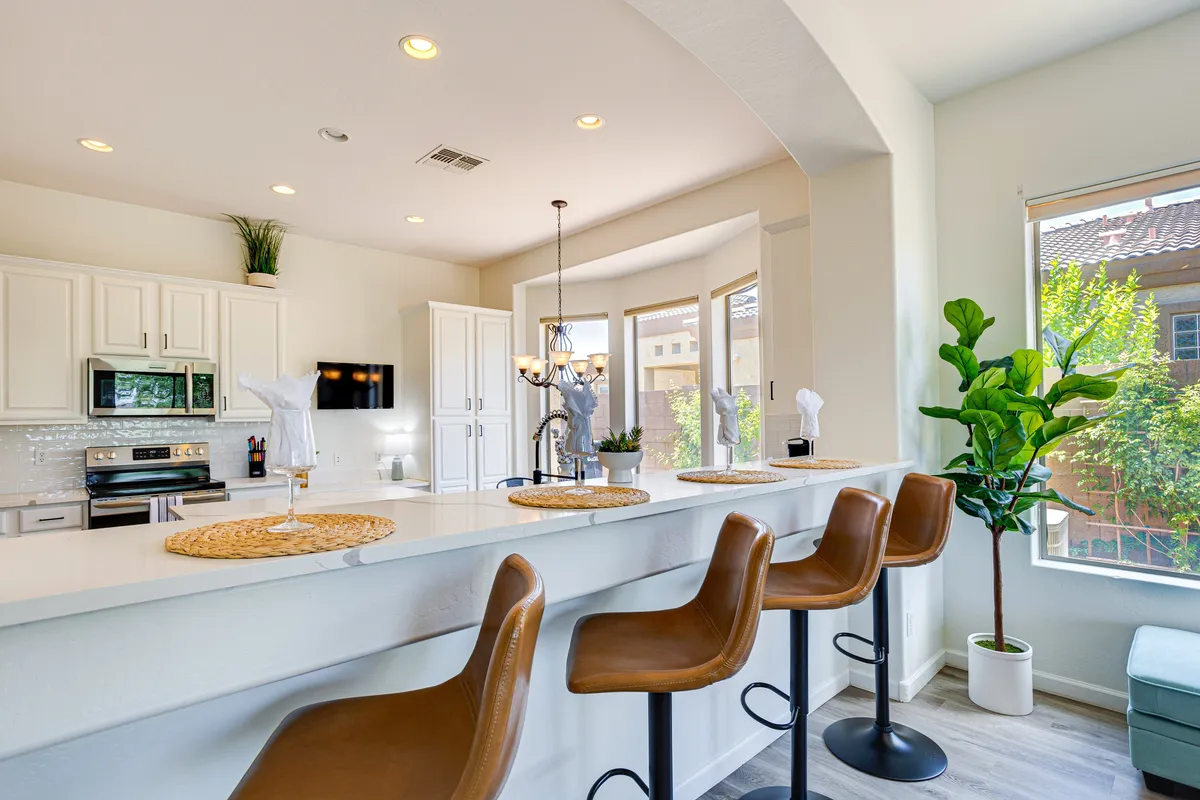A modern kitchen island with bar stools sits under a statement chandelier, adjacent to a large window that lets in natural light. The space is set for casual dining and gatherings, inviting shared meals and conversation.