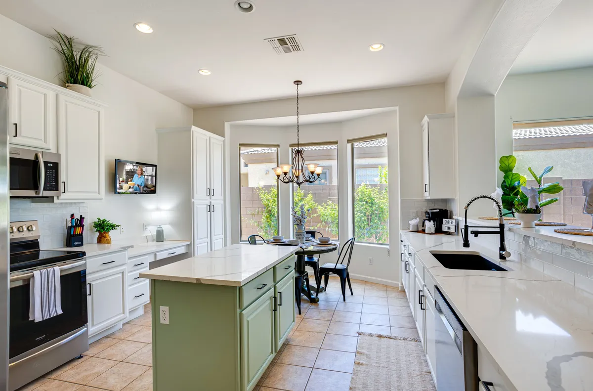A kitchen and dining area with a round table sits beside large windows that showcase lush greenery outside. The layout encourages gatherings for family meals, celebrations, and entertaining against a cheerful backdrop.