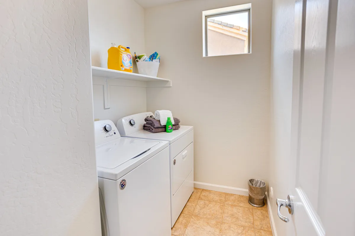A bright, tidy laundry room with a washing machine, drying machine, organized shelf with laundry supplies, and a window letting in natural light. The clean, functional layout highlights the ease of handling laundry tasks during any stay.